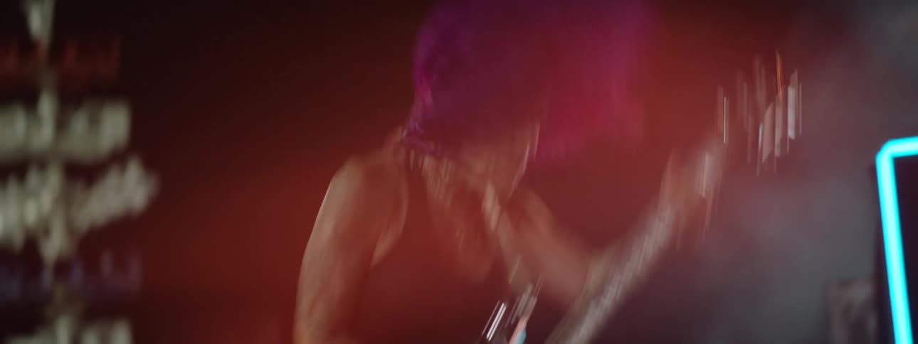 A musician plays an electric guitar vigorously on stage the person wears a dark shirt and has bright pink hair stage lights illuminate the area with orange and red colors