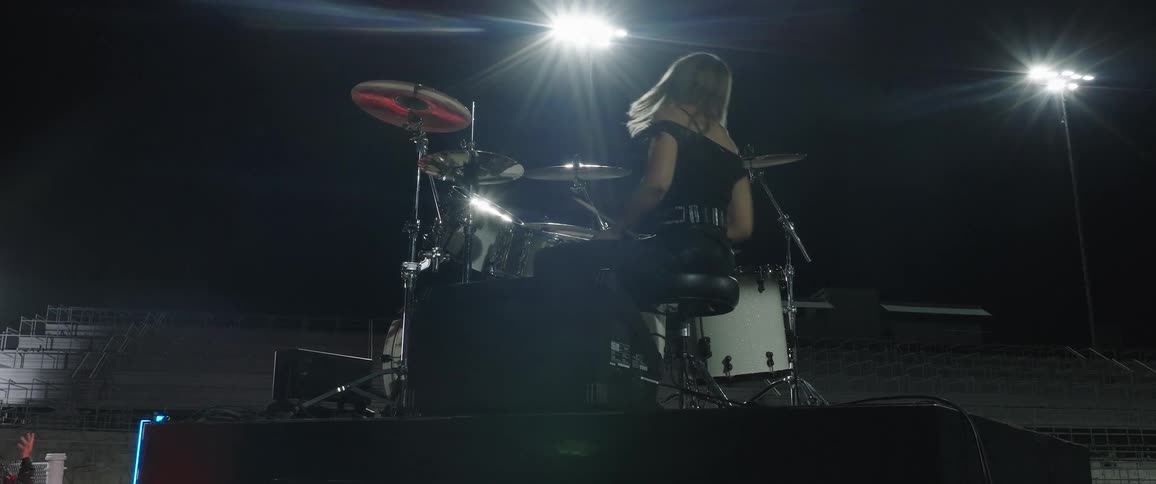 A low angle silhouette shows a long haired woman vigorously playing a white drum kit on a dark outdoor stage illuminated by a single bright spotlight from behind