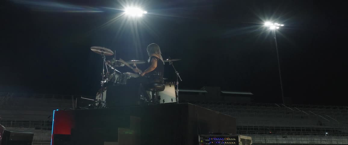A woman with long hair is shown from behind and below sitting on a drum kit and playing intensely on a large stage outdoors at night intensely illuminated by bright stadium lights