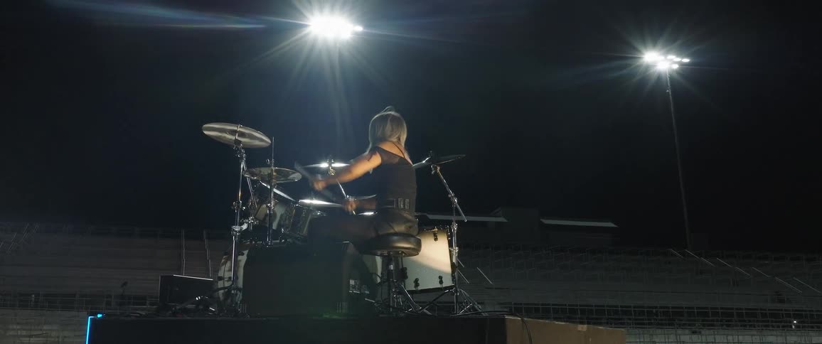 The video is a lowangle shot of a drummer silhouetted against bright stadium lights while playing a drum kit outdoors at night the drummers hair is flying as they hit the cymbals and the scene is framed by two powerful stadium lights in the bac