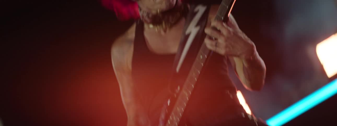Woman with purple hair plays guitar on a stage surrounded by light and smoke