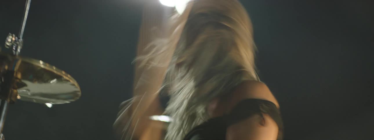 A low dynamic view shows a long haired musician in dark clothing playing a cymbal from a drum set on a dark outdoor stage illuminated by bright overhead floodlights