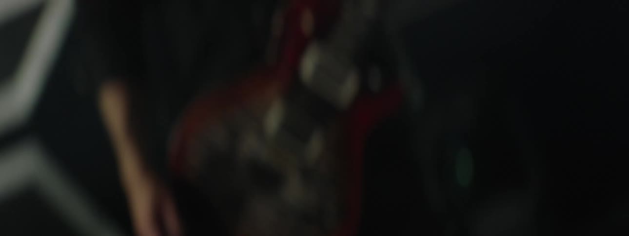A close low light view of a musicians hands shows him aggressively strumming a red and black electric guitar with a flame top finish on a dark stage