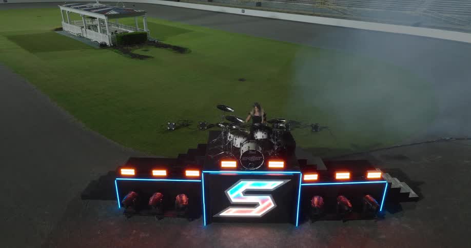 An elevated wide shot shows a four piece rock band on a lighted black stage with a large s logo and a drum set positioned on a race track or stadium surface next to a large green lawn with a small wooden structure