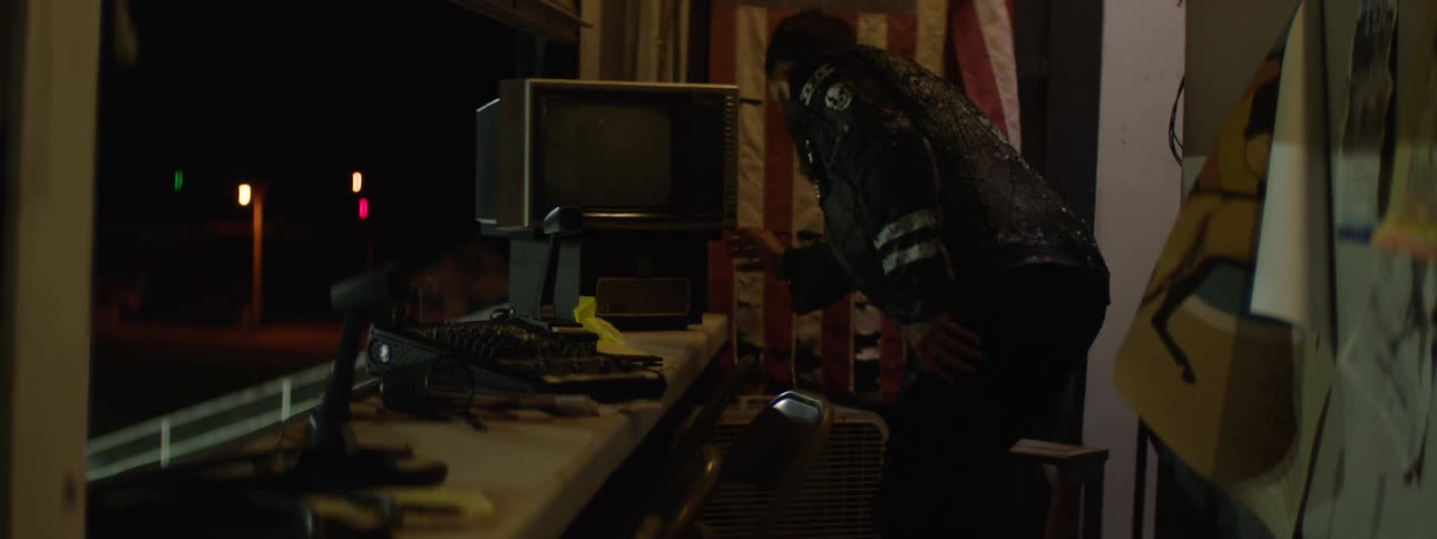 A person stands near a desk leans toward a monitor with a keyboard and fan on the desk indoors at night with light sources visible outside