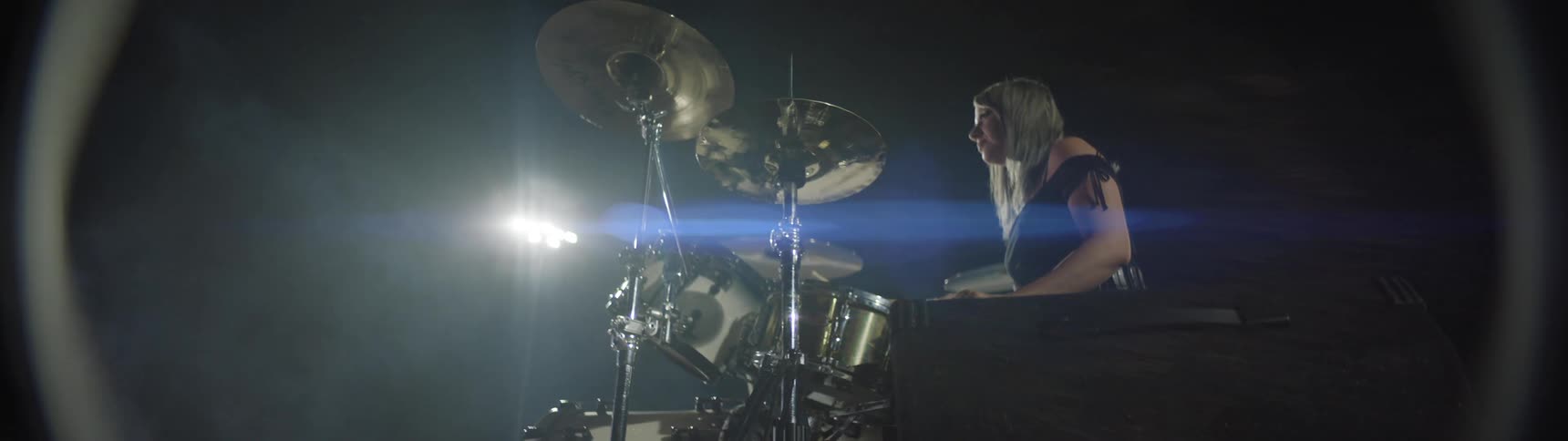 A woman with long blonde hair wearing a dark top is seen from the side as she sit sin front of a drum set with prominent cymbals against a dark background with bright stage lights creating flares and a smokey atmosphere