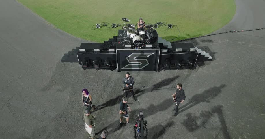 An elevated wide view shows a five person rock band on a black stage with a large s logo and a drum set surrounded by a film crew on a flat grey surface next to a large green lawn