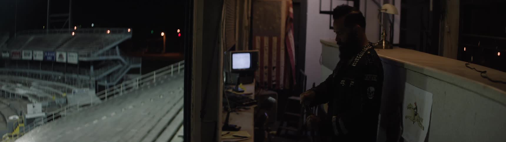 A bearded person stands in the shadow of an open doorway the person adjusts their hand on an object a bright monitor and a flag are visible inside and the outdoor scenario is a stadium