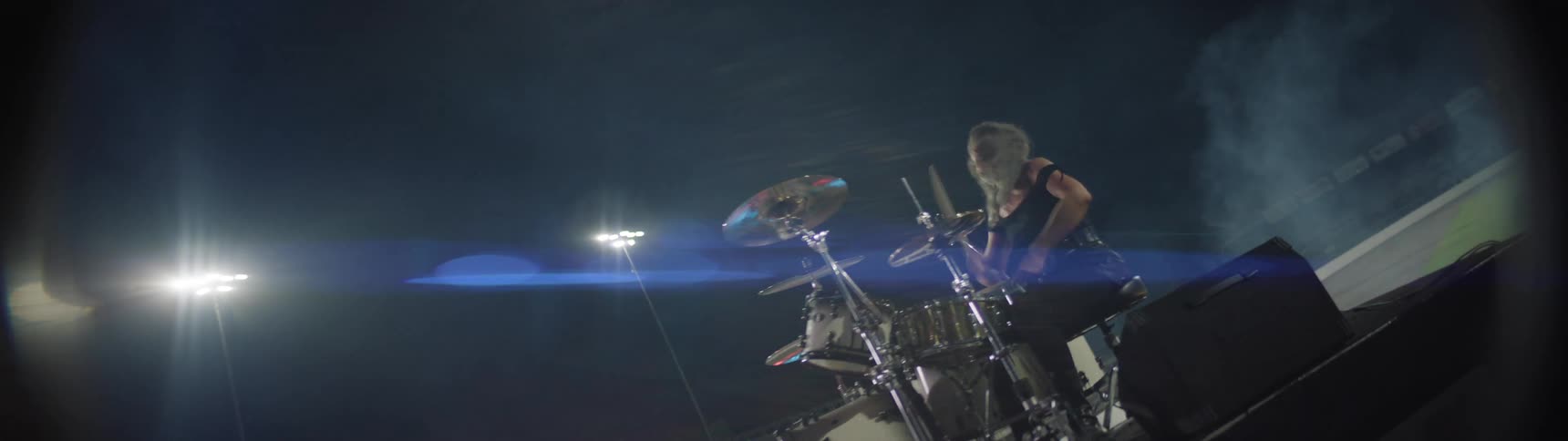 Drummer performs on lit stadium stage at night surrounded by haze and cinematic atmosphere