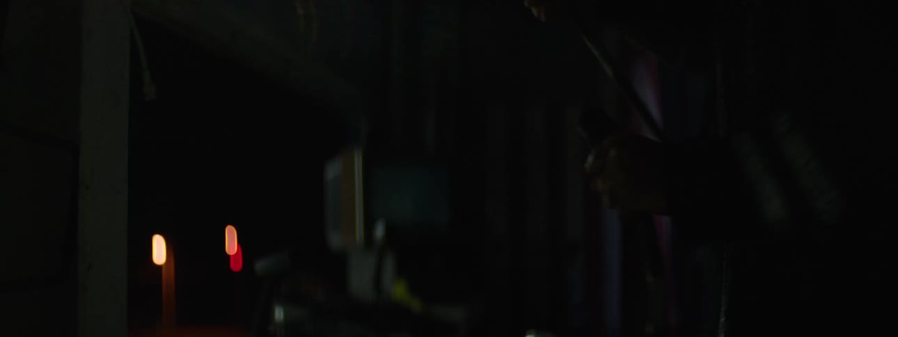 A man wearing dark clothing with reflective stripes unplugs a cable seen in a close view in a dimly lit space an out of focus tv monitor plunging the room in darkness