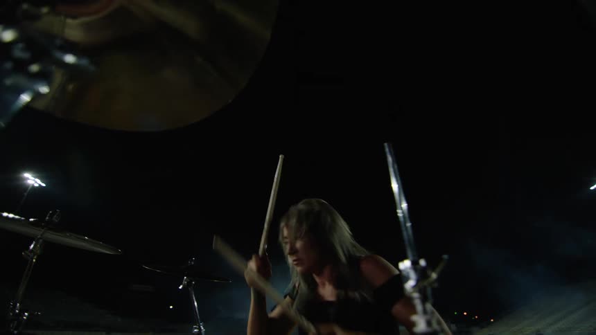 A dynamic low angle close view from inside the drum set shows a woman with long hair energetically striking the drums and cymbals with drumsticks during a nighttime outdoor performance
