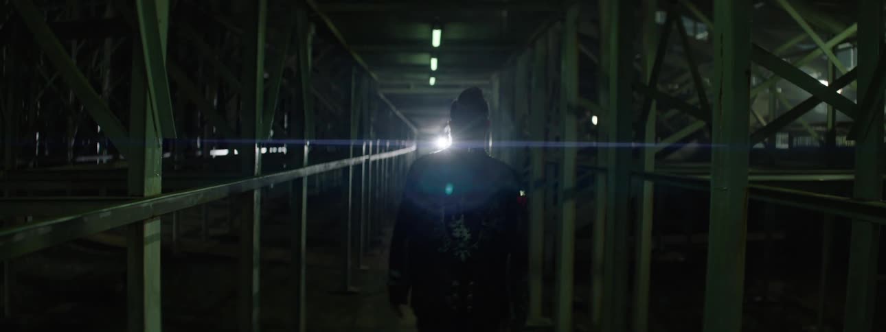 A low light shot from behind shows a person with a mohawk and a studded dark jacket walking down a long dark industrial corridor with overhead fluorescent lights and metal scaffolding on either side