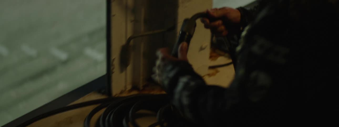 A close dark view shows a coil of black cable on a dirty window ledge until a person wearing a studded dark jacket with a skull patch reaches in from the right and handles a connector on the cable