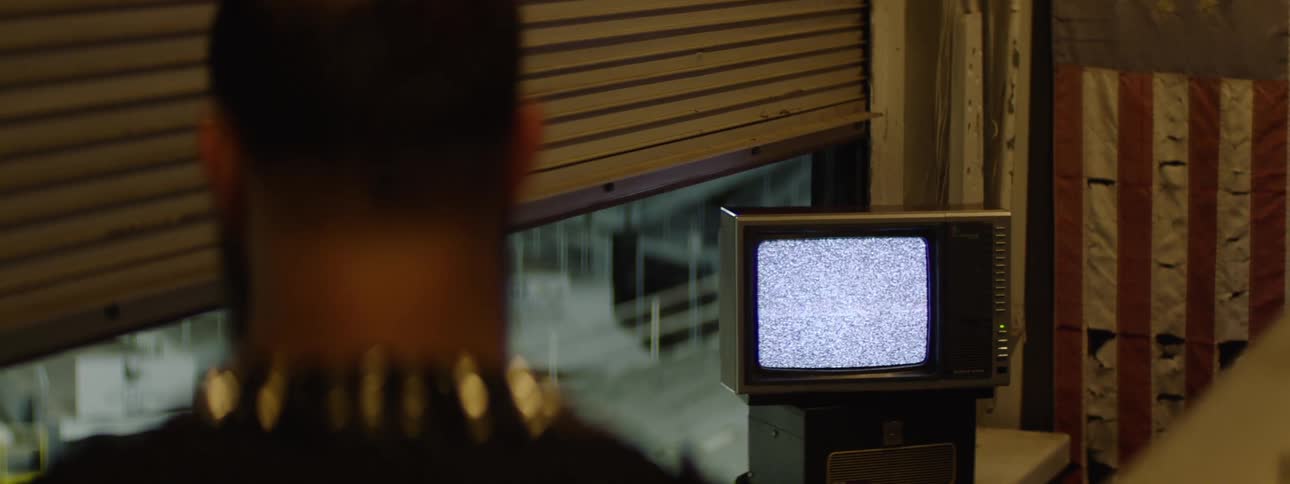 A close view from behind a man wearing a black garment with gold studs around the collar shows him looking toward a small vintage television set displaying static next to a window with closed blinds at night