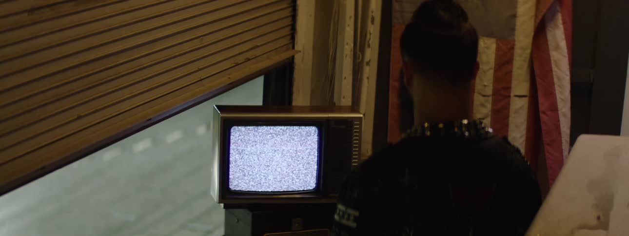 A dark view shows a person wearing a garment with silver studs around the collar looking at a small vintage television set displaying bright static positioned near an open window with light colored slatted shutters and a faint red and white striped flag