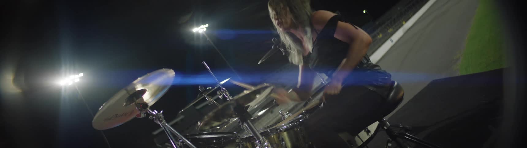 A person sits and plays a drum set hits the drums and cymbals with sticks the scene is a stadium at night with bright spotlights