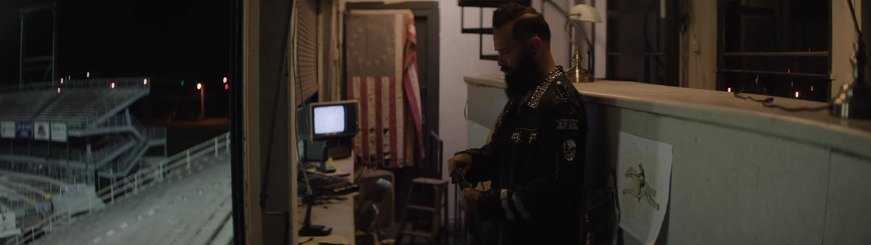 A bearded man with a mohawk wears a dark jacket covered in metal studs and skull patches turns off the lights of a stadium at night