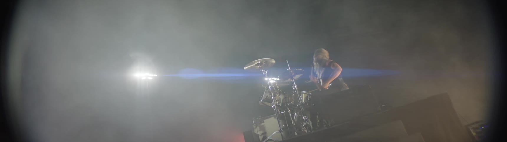 A female musician hits a drum set and cymbals with sticks on a stage surrounded by bright lights and fog during an outdoor music performance