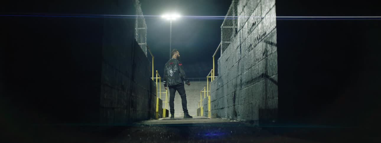 A young man in a black jacket stands at the entrance of a stadium at night illuminated by bright lights