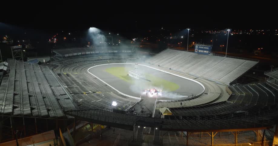 An empty stadium with large tiered seating is illuminated by tall lights at night a group of people and equipment are positioned on the oval track a structure stands on the track and smoke rises from the ground near the structure