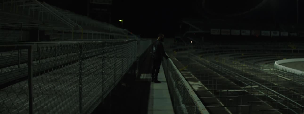 Person walks alone along a fenced walkway in an empty stadium at night