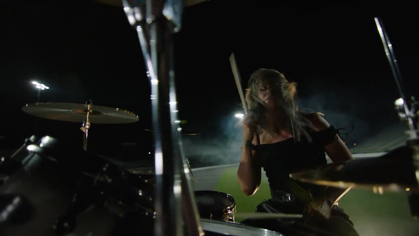 Woman plays drums energetically on a stage set up in a lit field at night