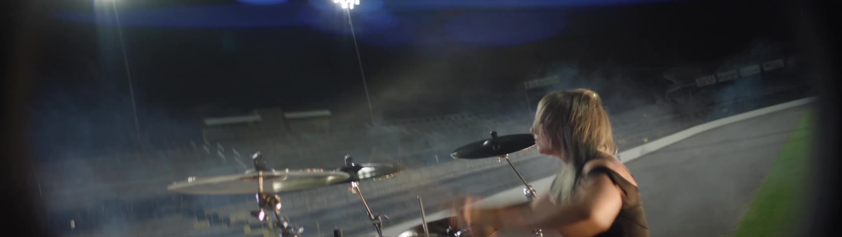 A person sits at a drum set hits the drums and cymbals with sticks performs on a stage the background is dark bright lights illuminate the musician from the side and behind