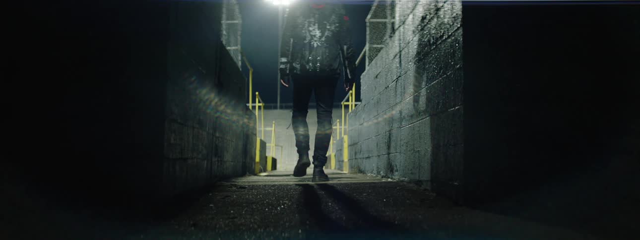 A low angle long view tracks a man wearing a dark leather jacket and pants as he walks away down a wet dark outdoor concrete alley illuminated by a single overhead light