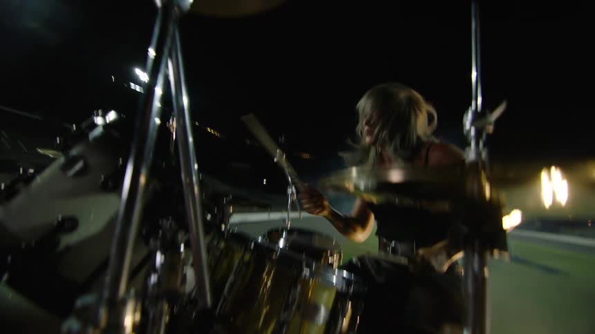 A dynamic low angle close view from the drum set shows a woman with long blonde hair in a dark top energetically striking the drums and cymbals with drumsticks during a nighttime stadium performance