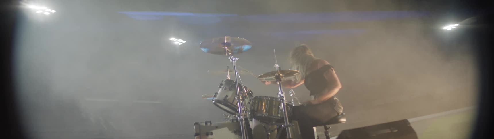 The video shows a female drummer performing live on a stage which is elevated and obscured by heavy stage fog or smoke she is energetically playing her silver drum kit with bright lights occasionally catching the cymbals