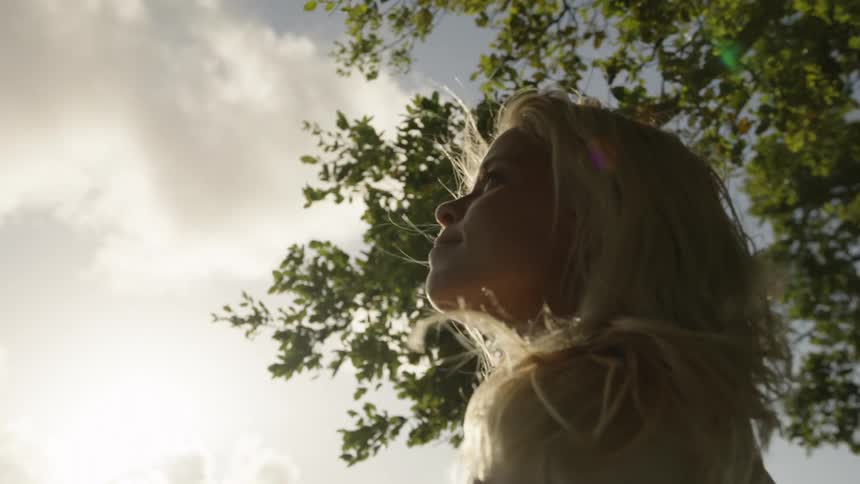 A persons head is viewed from the side and below the person looks upward toward a bright sky tree branches and leaves frame the top of the shot light shines from behind and illuminates the persons profile