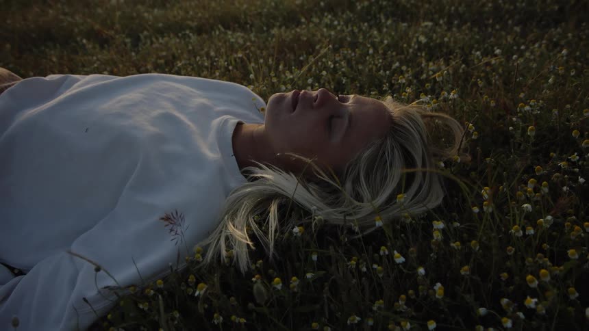 A blonde woman wearing a white sweatshirt lies relaxed in a field of small white and yellow flowers and dark grass as the setting sun illuminates her hair