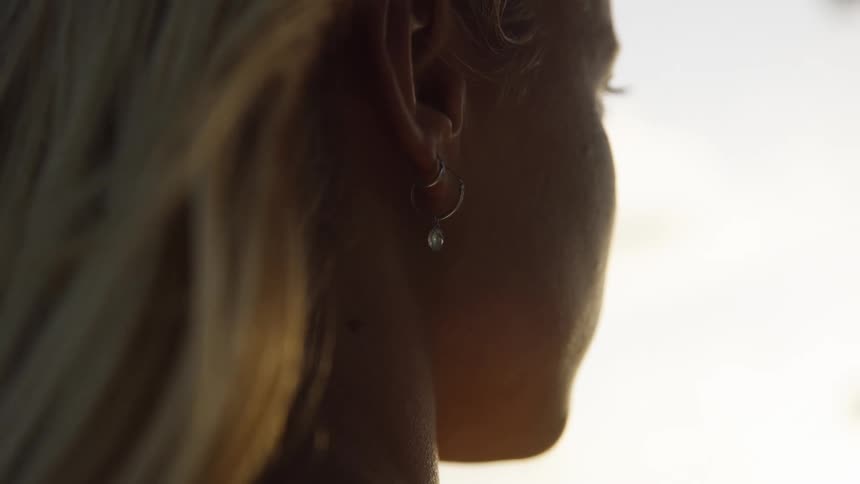 A young woman wearing earrings stands outdoors as light wind blows on her blonde hair