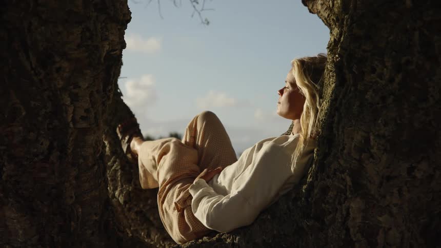 A side view of a young woman with blonde hair wearing a white longsleeved shirt and tan pants is shown resting comfortably within the cradle of a large tree fork looking out toward the blue sky and ocean on a sunny day
