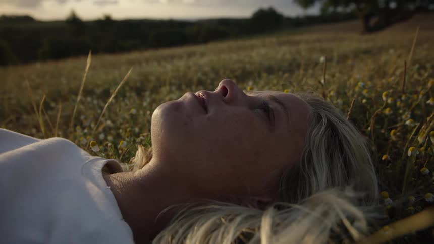 A closeup shot shows a blonde woman lying peacefully on her back in a field of tall grass and small white wildflowers her eyes are closed and the background is a rolling countryside landscape bathed in the warm golden light of sunset or sunrise