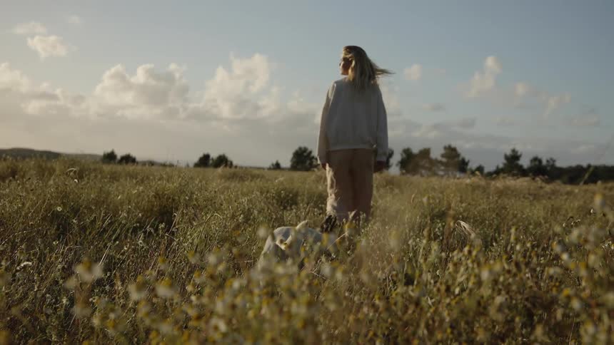 A blonde woman wearing a light colored sweatshirt and pants walks slowly away from the camera through a dry field of tall grass and small flowers as a small white dog follows her under a bright cloudy sky