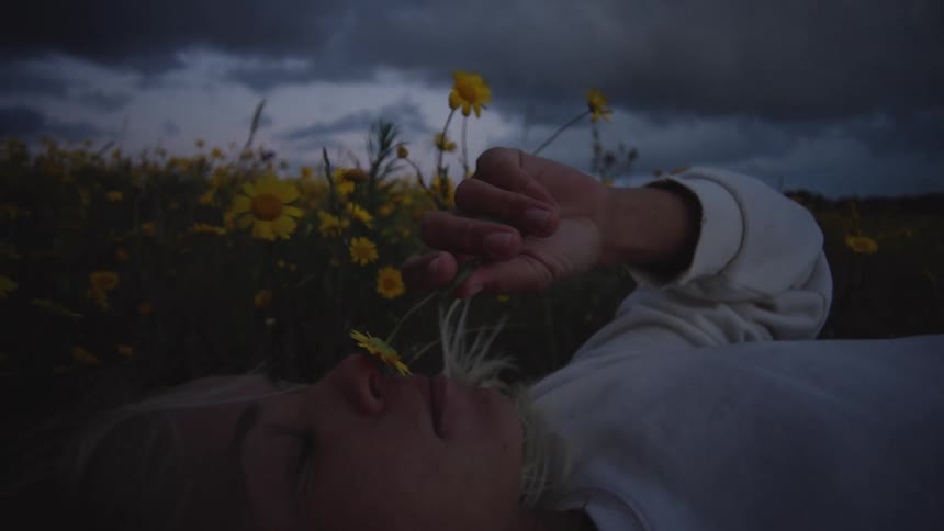 A low angle shot shows a young woman with blonde hair lying on her back in a dark field of yellow wildflowers gently holding one flower up to her face against a moody overcast sky