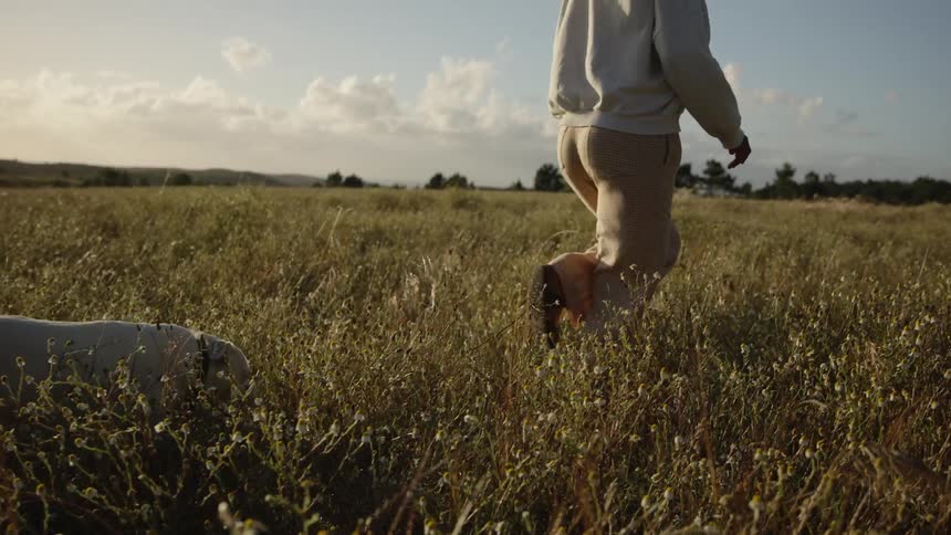 A person stands then walks away into a field a dog moves near the person tall grass and flowers cover the ground the sky is visible above