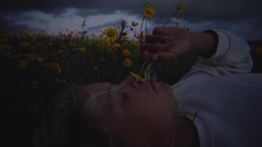 Woman lying in a field of yellow flowers holds one close to her face at dusk