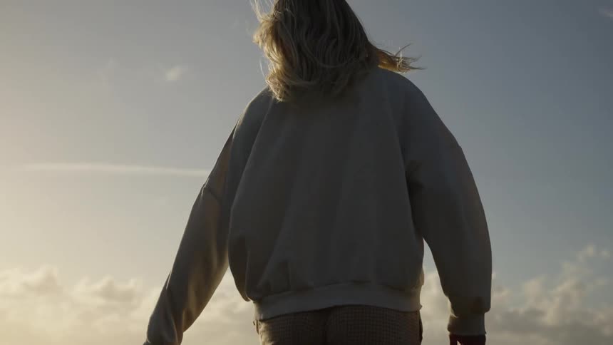 A medium shot from behind shows a blonde woman wearing a white sweatshirt and brown patterned pants walking slowly through a field of tall green grass and wildflowers against the bright evening sun