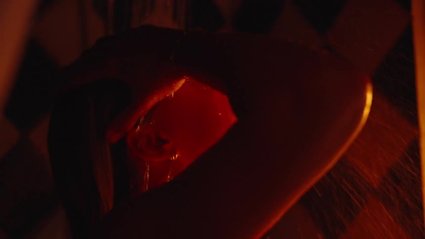 A close view shows a person taking a dark moody shower rubbing their hands over their face and wet head as water runs down their skin all illuminated by warm intense orange light against a dark patterned tile wall