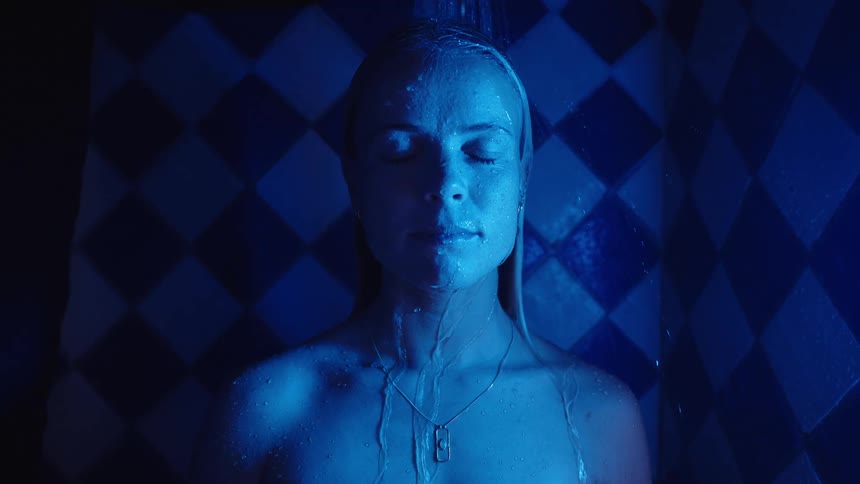 A woman stands in a shower under intense blue light closing her eyes as water streams over her against a checkered wall