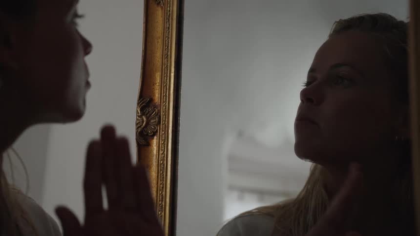 Woman looks at her reflection in the mirror touching her face with a pensive expression