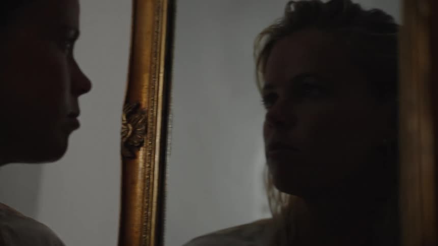 Blonde woman stares at her reflection in a gilded mirror