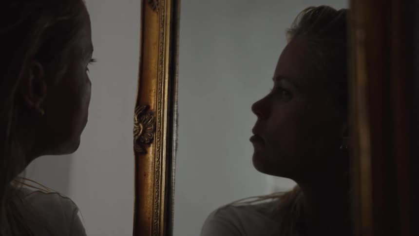 A womans profile faces the ornate frame of a mirror while her reflection faces the camera all set against a dark indoor backgroun