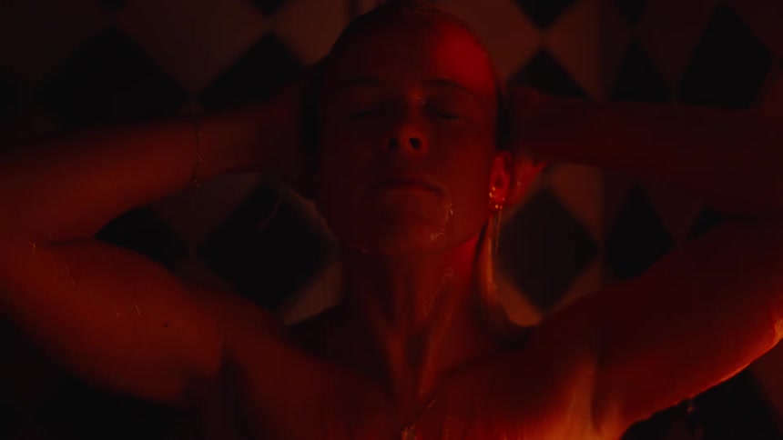 A close moody shot shows a blonde woman in a dark shower washing her hair under running water her body illuminated by dramatic warm red light against a background of dark diamond patterned tiles