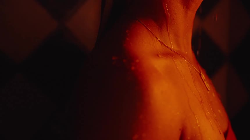 A close intimate view of a womans neck and side shows beads of water running down her skin illuminated by dramatic warm orange light against a dark tiled background