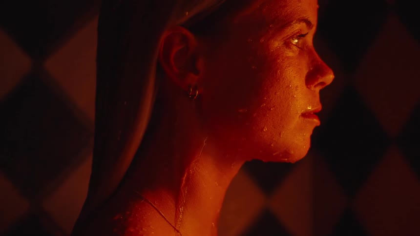 A closeup side view shows a womans face and neck water runs down the skin surface the scene is illuminated by a warm orange light source and a diamond pattern is visible on the dark background wall