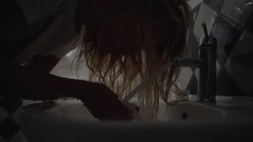 A blonde woman in a white shirt turns on a chrome faucet and bends over a white sink to wash her face with running water in a dark bathroom with blue and white diamond patterned tiles then looks up at a gold framed mirror