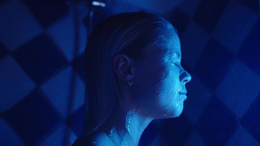 A person stands under running water the persons head and shoulders are visible a hand touches the head water streams down the face and hair and blue light illuminates the person against a tiled wall